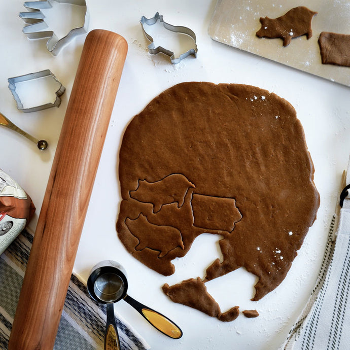 baker's rolling pin on tabletop with rolled out cookie dough in the shape of pigs and states